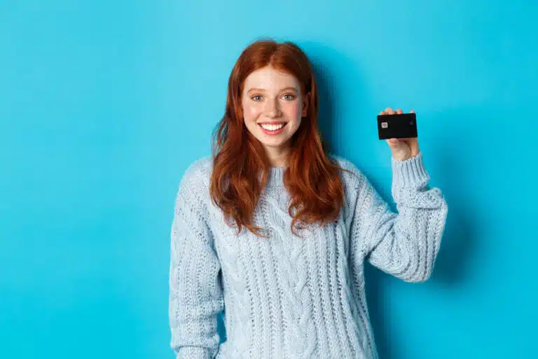 cute redhead girl in sweater showing credit card, smiling at camera, standing over blue background