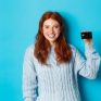 cute redhead girl in sweater showing credit card, smiling at camera, standing over blue background